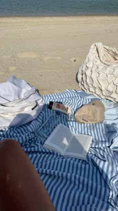 an open book on a blue and white striped blanket next to the beach with other items