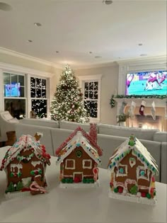 three gingerbread houses are on a table in front of a christmas tree and television