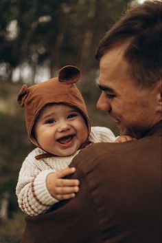 a man holding a baby wearing a brown bear hat and smiling at the camera with trees in the background