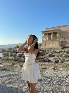 a woman in a white dress is talking on her cell phone near the ruins of an ancient city