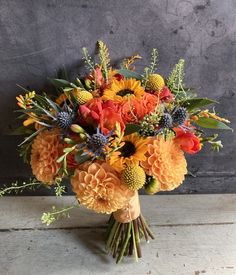 a bouquet of orange and yellow flowers sitting on top of a wooden table next to a gray wall