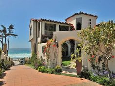 a white house with lots of flowers on the front and side of it next to the ocean