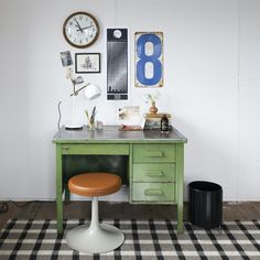 a green desk with a clock on the wall above it and an orange stool in front of it