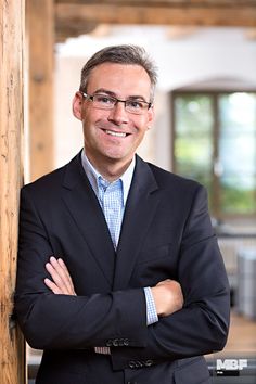 a man wearing glasses and a suit standing in front of a wooden wall with his arms crossed