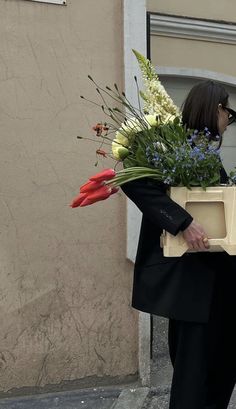 a woman carrying a box with flowers in it on the sidewalk next to a building