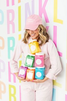 a woman in pink is holding up some colorful cups and smiling at the camera while wearing a pink hat