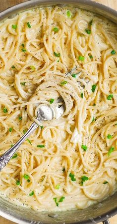 a bowl filled with noodles and peas on top of a wooden table next to a silver spoon