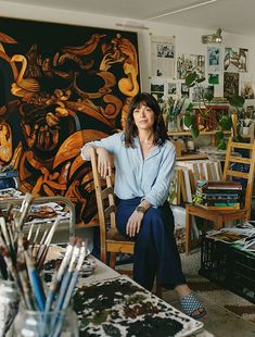 a woman sitting on a chair in front of a table with many paintings and brushes