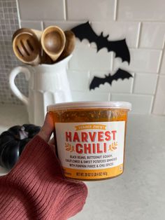 a person holding up a jar of harvest chilli in front of a kitchen counter