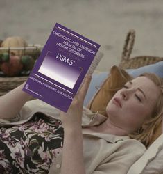 a woman laying on a couch holding up a book in front of her face and looking at the camera