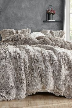 a bed with grey fur on it in a room next to a window and wooden floor