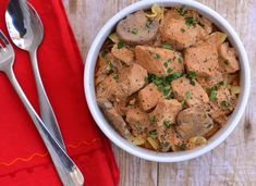 a white bowl filled with meat and pasta next to a spoon on top of a red napkin