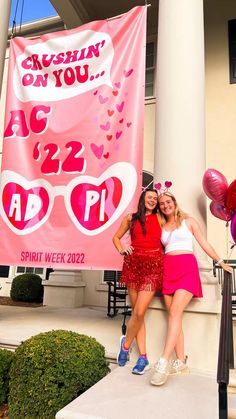 two women standing next to each other in front of a pink sign with hearts on it