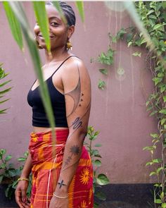 a woman standing in front of a plant with tattoos on her arm and chest, wearing an orange skirt