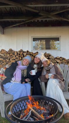 two women sitting on chairs next to a fire pit with logs in the background and one woman holding a mug