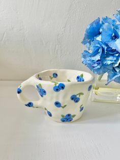 blue flowers are in a vase next to a white cup with water on the table