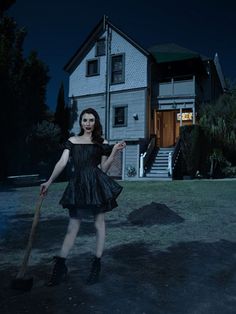 a woman in a black dress holding a broom and standing in front of a house at night