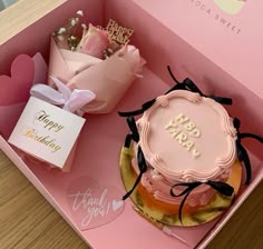 a pink birthday cake in a pink box with some flowers and cards on the table