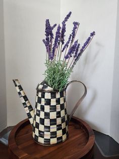 a checkered watering can with lavenders in it sitting on a wooden tray next to a white wall