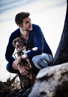 a man holding a small dog in his lap while sitting on a tree stump with the background of a rock and trees