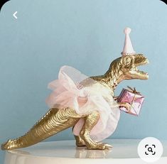 a gold dinosaur figurine in a tutu skirt and party hat holding a present