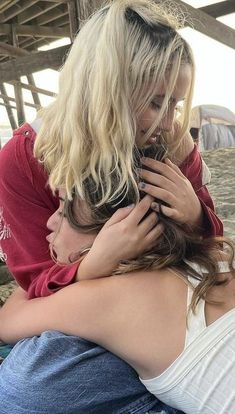 two women hug each other while sitting on the ground in front of an ocean pier