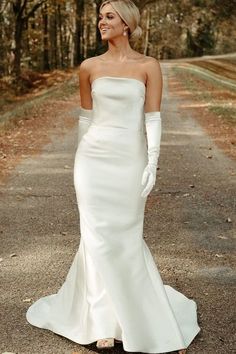 a woman in a white dress and gloves standing on the side of a dirt road