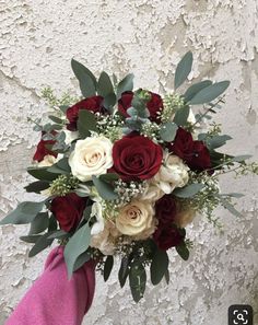 a bridal bouquet with roses and greenery in someone's hand against a white wall