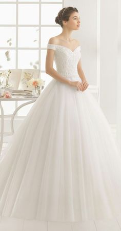 a woman in a white wedding dress standing by a window with flowers on the table