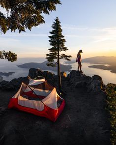 a person standing on top of a mountain next to a tent and tree with the sun setting in the background