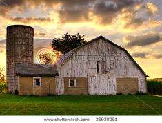 an old barn and silo with the sun setting in the sky behind it on a farm