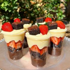 four dessert cups with strawberries and chocolate on top are sitting on a plate in front of some bushes