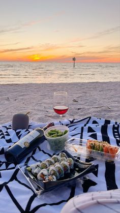 a plate of sushi and wine on the beach at sunset, with a glass of wine in the foreground