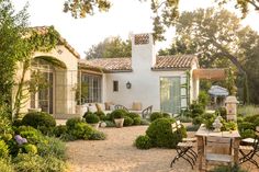 an outdoor patio with chairs and tables surrounded by greenery, shrubs and trees in front of a white stucco house