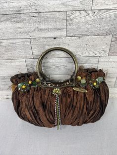 This elegant Mary Frances women's purse features a beautiful brown silk bag with intricate beaded flower accents. The bag has a magnetic closure and a top handle made of gold-tone metal. The exterior is made of soft silk in a rich brown color, while the interior is lined with brown satin. The bag is small in size, measuring 10.5 inches in width, 5.5 inches in height, and 4 inches in depth. It comes with a charming bag charm and is perfect for any occasion. This luxury purse is a must-have accessory for any fashion-conscious woman who wants to add a touch of luxury to her wardrobe. PR Room On wood shelves by window shelf 2 Mary Frances Olive Green Brown Handbag, Beaded Brown Tote Shoulder Bag, Brown Beaded Tote Shoulder Bag, Brown Beaded Handheld Bag, Handheld Brown Beaded Bag, Travel Brown Beaded Shoulder Bag, Brown Top Handle Evening Bag For Party, Beaded Brown Shoulder Bag For Travel, Bohemian Handheld Embellished Bag