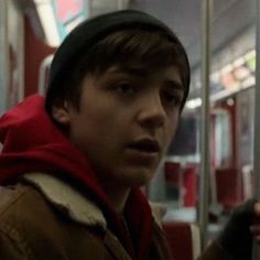 a young man wearing a hat and jacket on a subway train looking at the camera