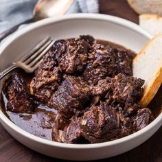 a bowl filled with meat and bread on top of a wooden table next to a fork