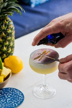 a person holding a spoon in front of a glass with a drink on it next to pineapples and other fruit