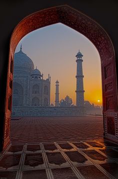 the sun is setting in front of an arch leading to a building with two towers