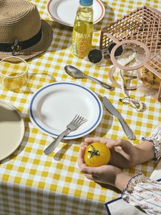 a person holding an orange on a yellow and white checkered table cloth with silverware