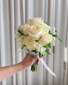 a person holding a bouquet of white roses and greenery in front of a curtain