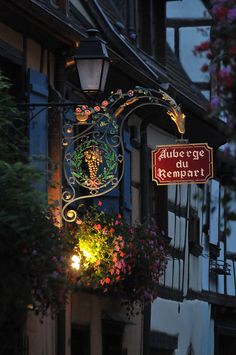 an illuminated sign hanging from the side of a building with flowers growing out of it