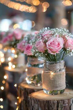 three mason jars with pink roses in them on a tree stump, surrounded by string lights