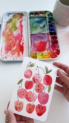 someone is holding up a watercolor painting in front of two trays of paint