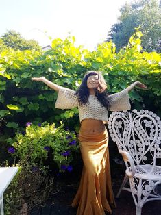 a woman standing next to a white bench in front of some bushes and flowers with her arms outstretched