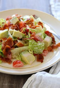 a salad with bacon, lettuce and tomatoes on a white plate next to a fork