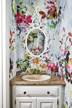 a white sink sitting under a mirror next to a wall covered in floral wallpaper