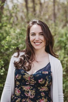 a smiling woman in a floral dress and cardigan standing in front of some trees