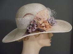 "Wide brim straw with a bouquet of flowers and veiling. Perfect for a garden party or Kentucky Derby. Off white straw has a 2.5\" white embossed ribbon band at crown. Flowers in shades of beige/mauve/purple, lilac. Mauve ribbon detail with white veiling. Mauve biot feathers. Trimmed in white grosgrain ribbon. Millinery wired brim to hold shape. Fit 22.5\" to 23\"" Cream Cloche Hat With Curved Brim For Garden Party, Cream Wide Brim Cloche Hat For Garden Party, Wide Brim Fascinator With Handmade Flowers For Garden Party, Brimmed Cloche Hat For Kentucky Derby, Elegant Cloche Straw Hat For Garden Party, Curved Brim Hat With Handmade Flowers For Garden Party, Brimmed Cloche Hat For Kentucky Derby Vintage Events, Elegant Cream Sun Hat For Garden Party, Vintage Short Brim Fascinator For Garden Party