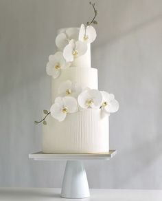 a three tiered cake with white flowers on top is sitting on a table in front of a gray wall